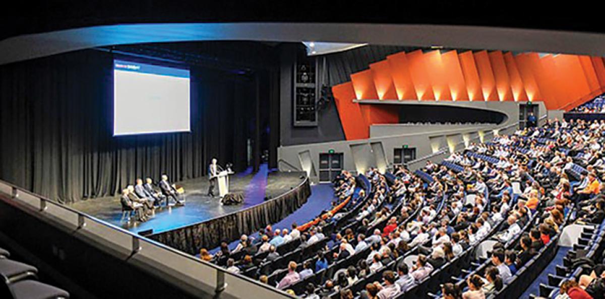 Side view of an auditorium filled with people. Someone is giving a speech and there is a screen behind them.