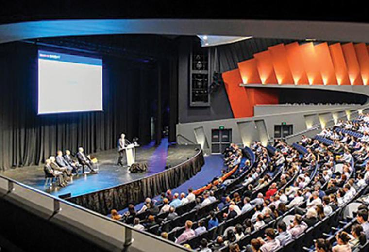 Side view of an auditorium filled with people. Someone is giving a speech and there is a screen behind them.